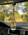A clear glass car air freshener bottle with a light wood cap and a tan rope hanger, hanging from a rearview mirror inside a car with a blurred green forest background visible through the windshield.