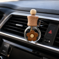 A car air freshener with a wooden top and a brown flower-shaped diffuser in a clear glass bottle, clipped onto a car dashboard air vent.